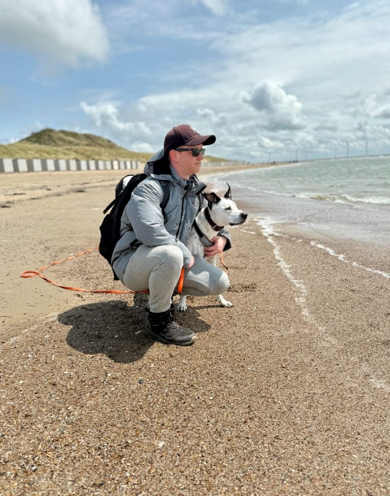 Hundeschule Partnerschnauze Mann kniet am Strand neben einem Hund und schaut aufs Wasser. Wolken am Himmel.