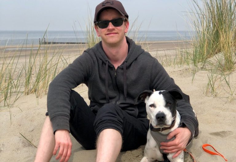 Hundetrainer Hauke Gehlen Mann sitzt am Strand mit seinem Hund, umgeben von Sand und Gras.